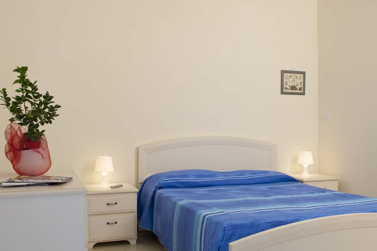 A bedroom with a bed, two bedside tables, a nightstand, a lamp, and a potted plant.