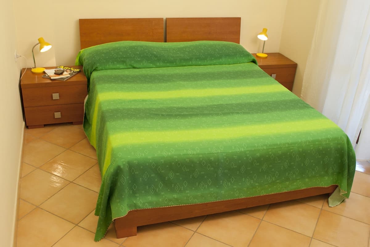 A neatly made bed with a green bedspread, two bedside tables with lamps, and a tiled floor.