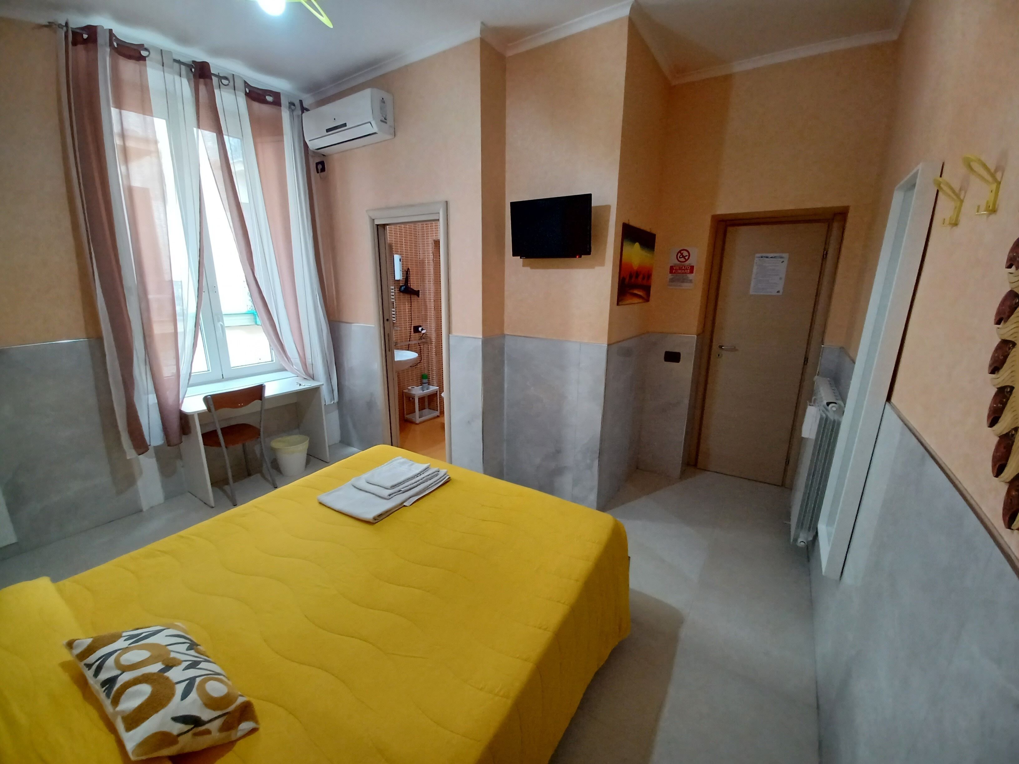 Ein Hotelzimmer mit einem gelben Bett, einem Schreibtisch, einem Stuhl und einem kleinen Tisch. Es gibt ein Fenster mit Vorhängen, eine Klimaanlage und eine Tür mit einem Schild.