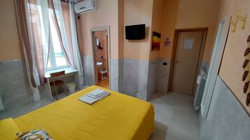 Ein Hotelzimmer mit einem gelben Bett, einem Schreibtisch, einem Stuhl und einem kleinen Tisch. Es gibt ein Fenster mit Vorhängen, eine Klimaanlage und eine Tür mit einem Schild.