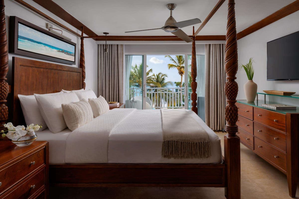 A spacious bedroom with a large bed, wooden headboard, and a balcony with a view of palm trees and the ocean.