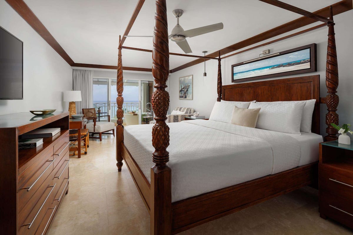 A spacious bedroom with a four-poster bed, a flat-screen TV, a desk with a lamp, and a balcony with a view.