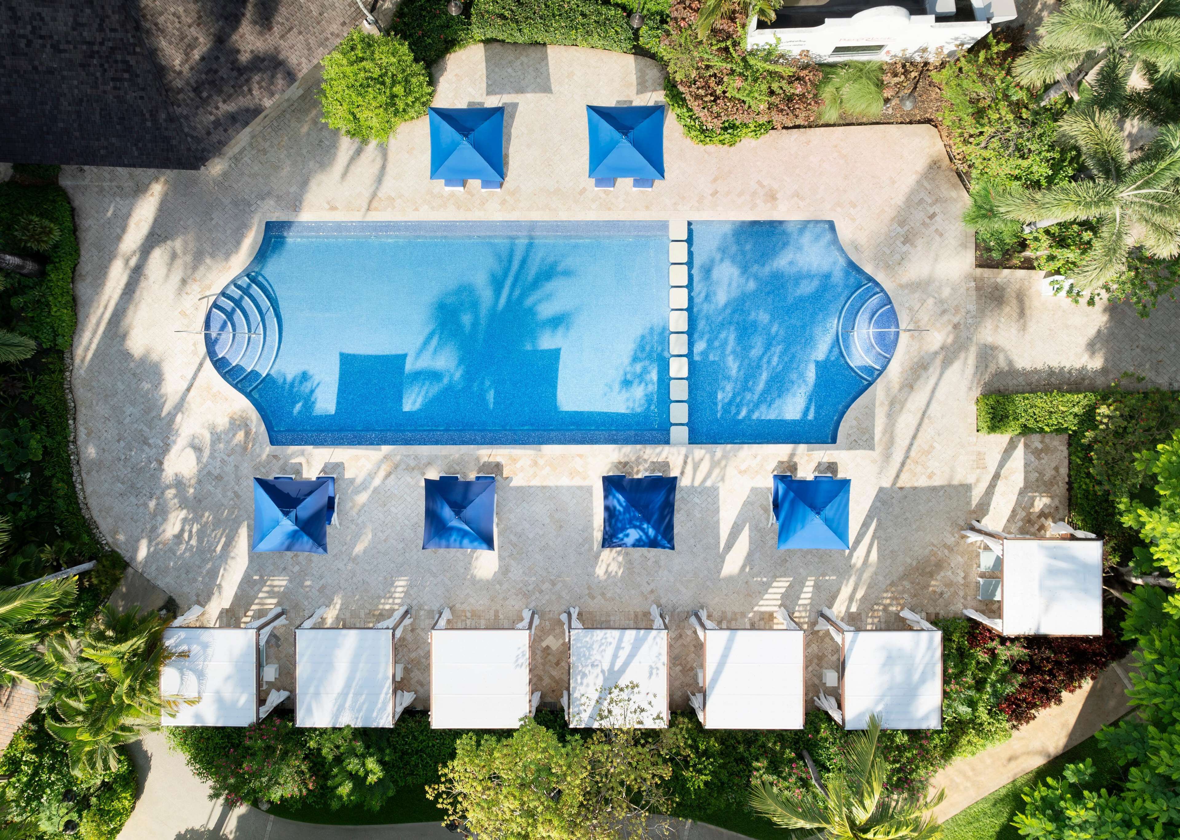 An aerial view of a swimming pool area with lounge chairs and umbrellas, surrounded by greenery and a paved walkway.