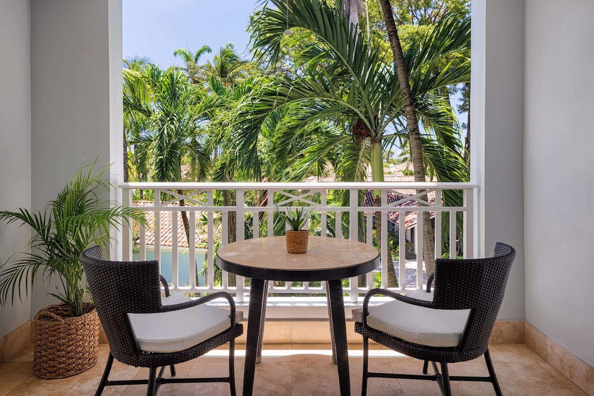 A balcony with a round table and two wicker chairs, surrounded by white railings and lush greenery.