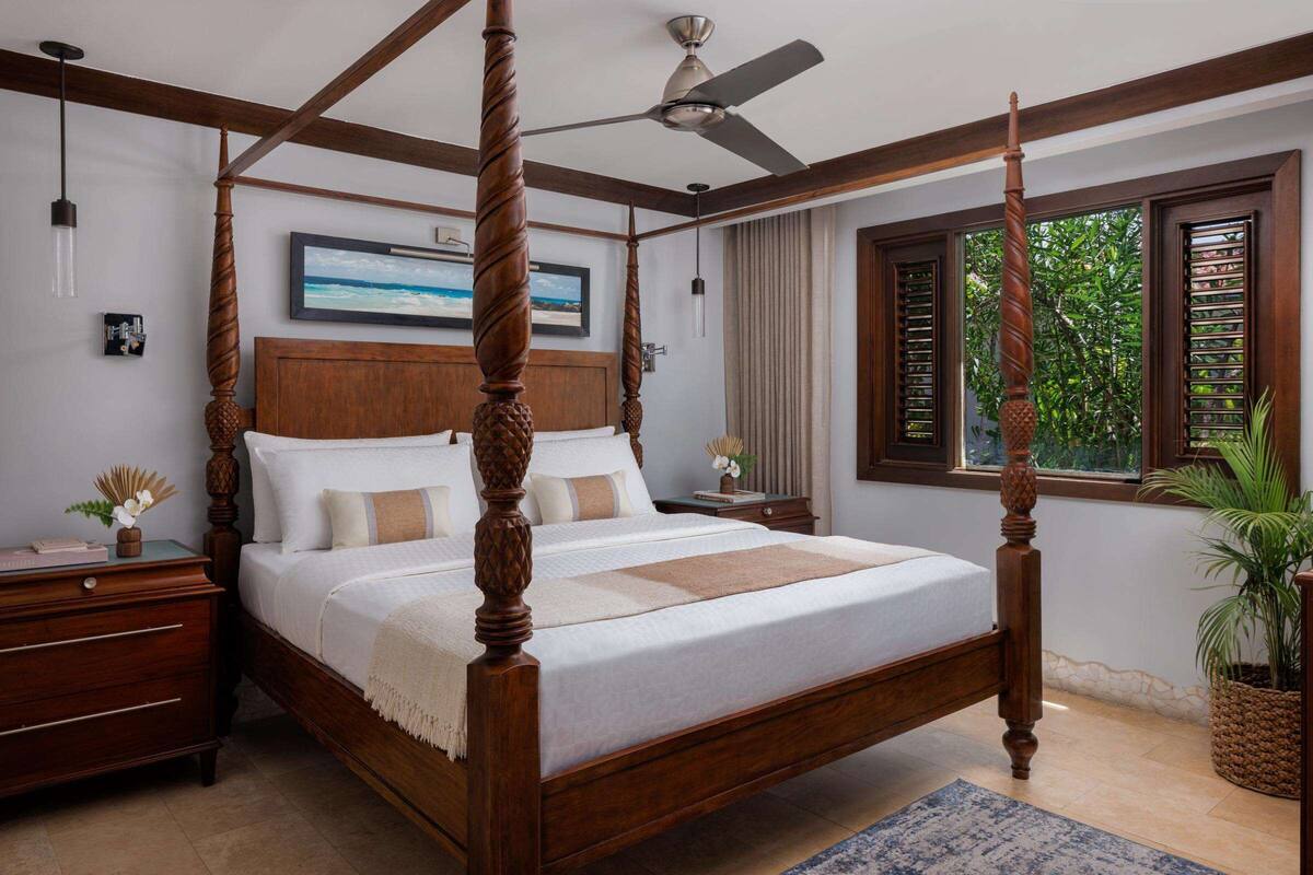 A four-poster bed with a wooden frame and a ceiling fan, a nightstand with a lamp, a framed picture on the wall, and a window with shutters.