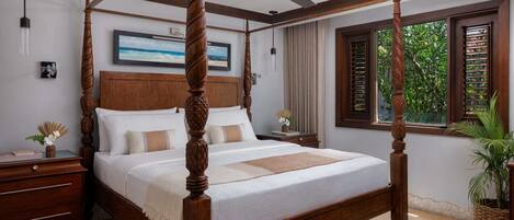 A four-poster bed with a wooden frame and a ceiling fan, a nightstand with a lamp, a framed picture on the wall, and a window with shutters.