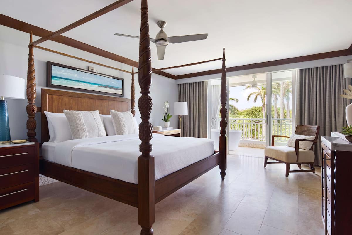 A four-poster bed with a ceiling fan, a large window with curtains, a desk with a chair, and a nightstand with a lamp.