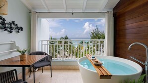A balcony with a view of palm trees and the sea, a wooden table with chairs, and a freestanding bathtub with a wooden tray. - Sandals Barbados - ALL INCLUSIVE Adults Only (Maxwell)