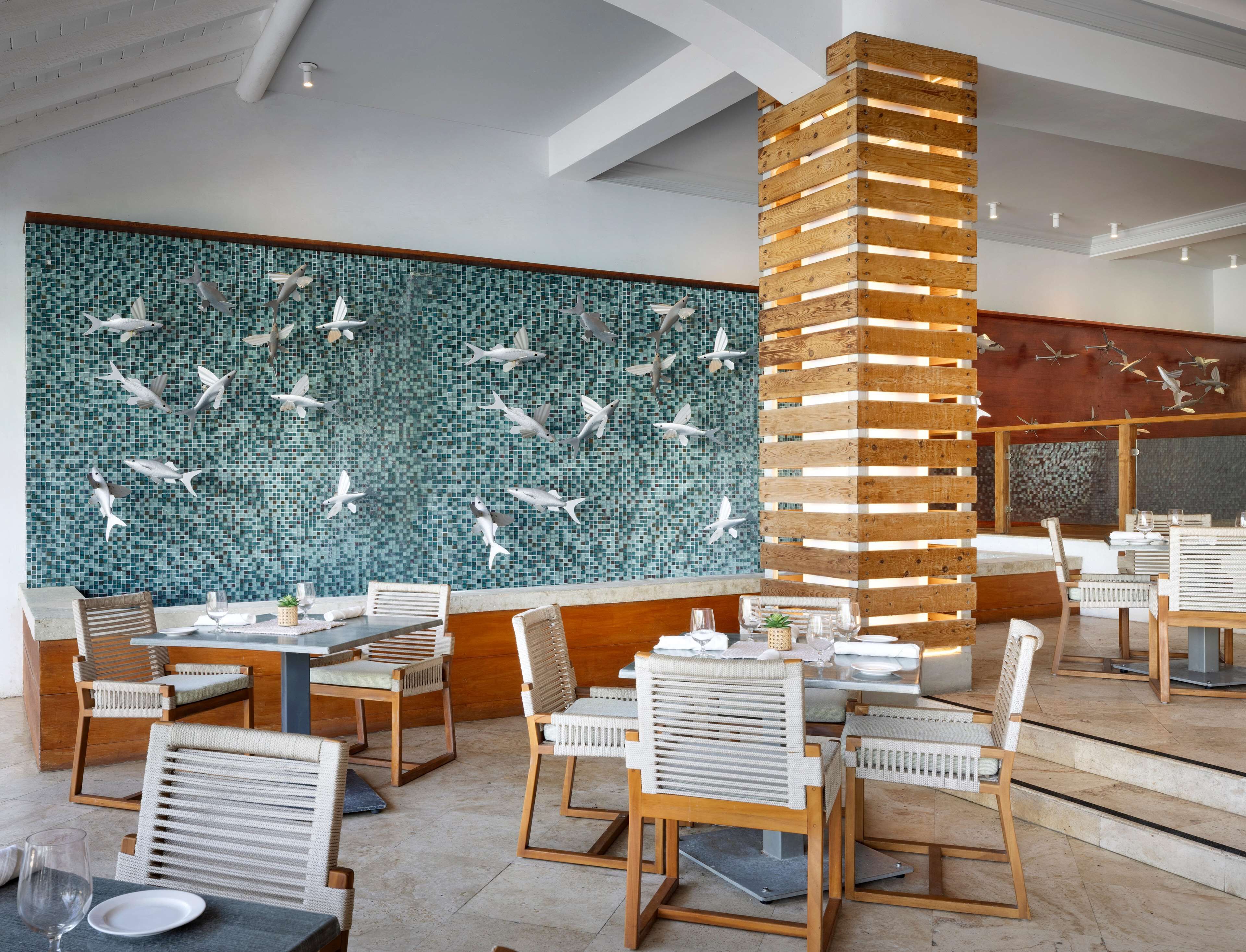 A restaurant with a mosaic wall featuring fish and birds, wooden chairs, and tables set for dining.