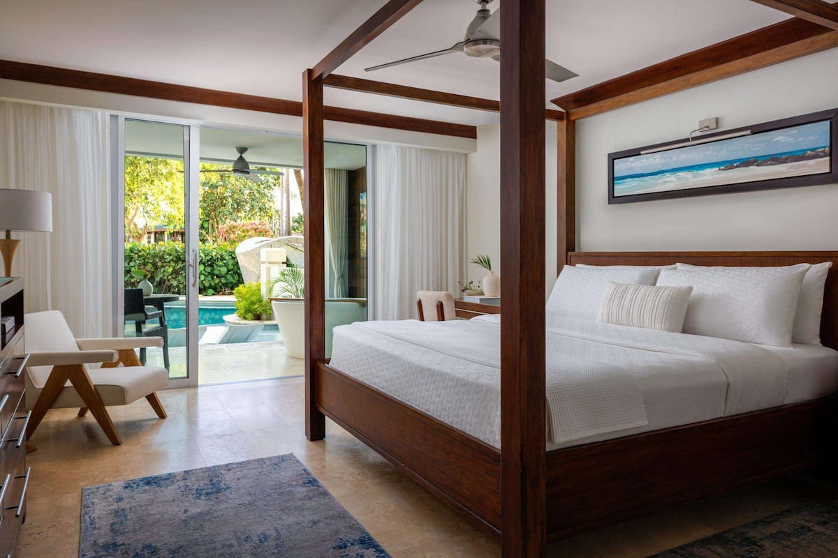 A bedroom with a four-poster bed, a view of a pool and greenery, and a ceiling fan.