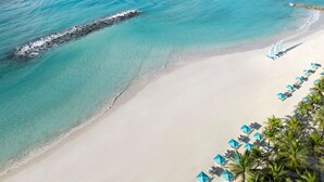An aerial view of a beach with clear turquoise water, a rocky outcrop, and a row of blue umbrellas and lounge chairs. - Sandals Barbados - ALL INCLUSIVE Adults Only (Maxwell)