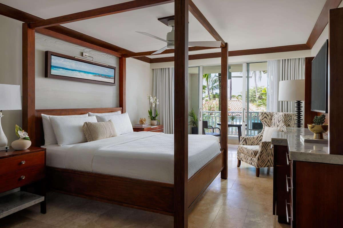 A four-poster bed with a wooden frame, a bedside table with a lamp, and a view of the outdoor balcony.