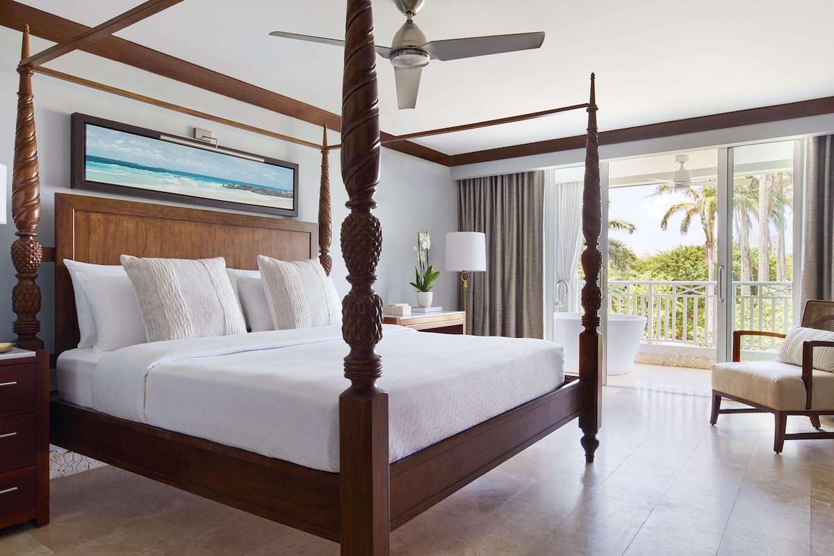 A four-poster bed with a ceiling fan, a large window with a view of palm trees, a chair, and a desk with a lamp.
