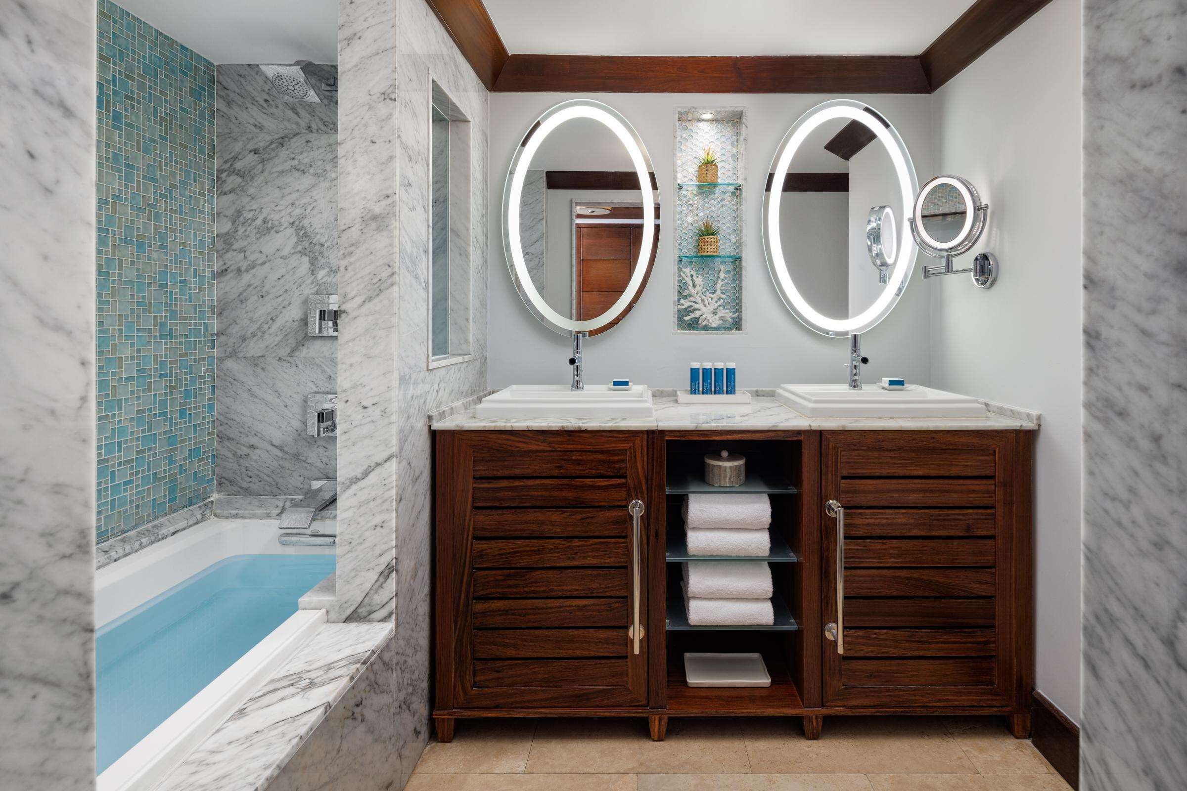 A bathroom with a marble wall, a wooden vanity with two sinks, and a built-in bathtub.