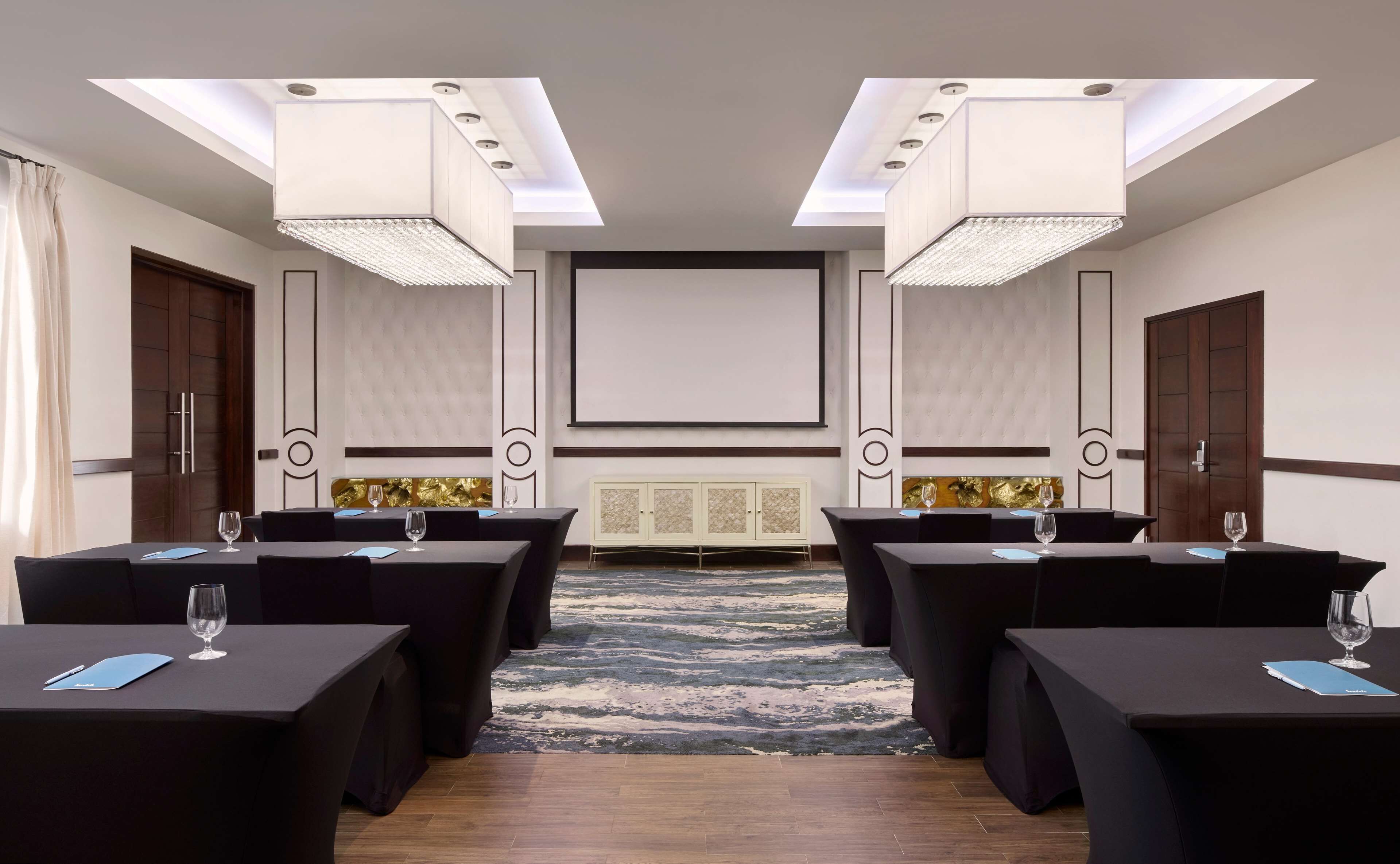 A conference room with a large screen, multiple tables with chairs, and a patterned carpet.