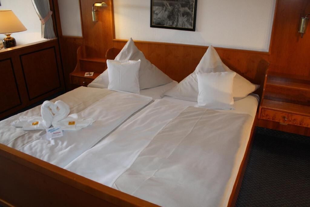 Habitación de hotel con una cama grande, dos almohadas, una toalla doblada, una mesita de noche, una lámpara y un cuadro en la pared.