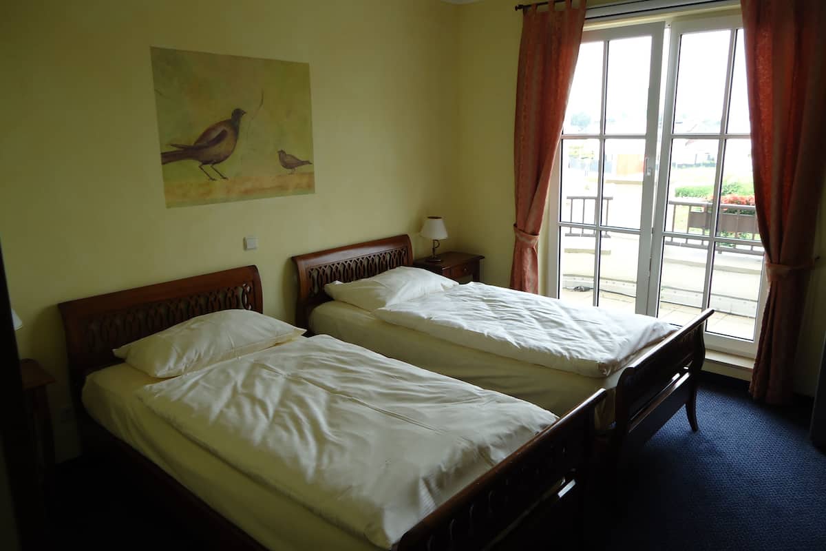 Ein Zimmer mit zwei Einzelbetten, einem Nachttisch mit Lampe, einem Vogelbild an der Wand und einem Fenster mit Vorhängen.