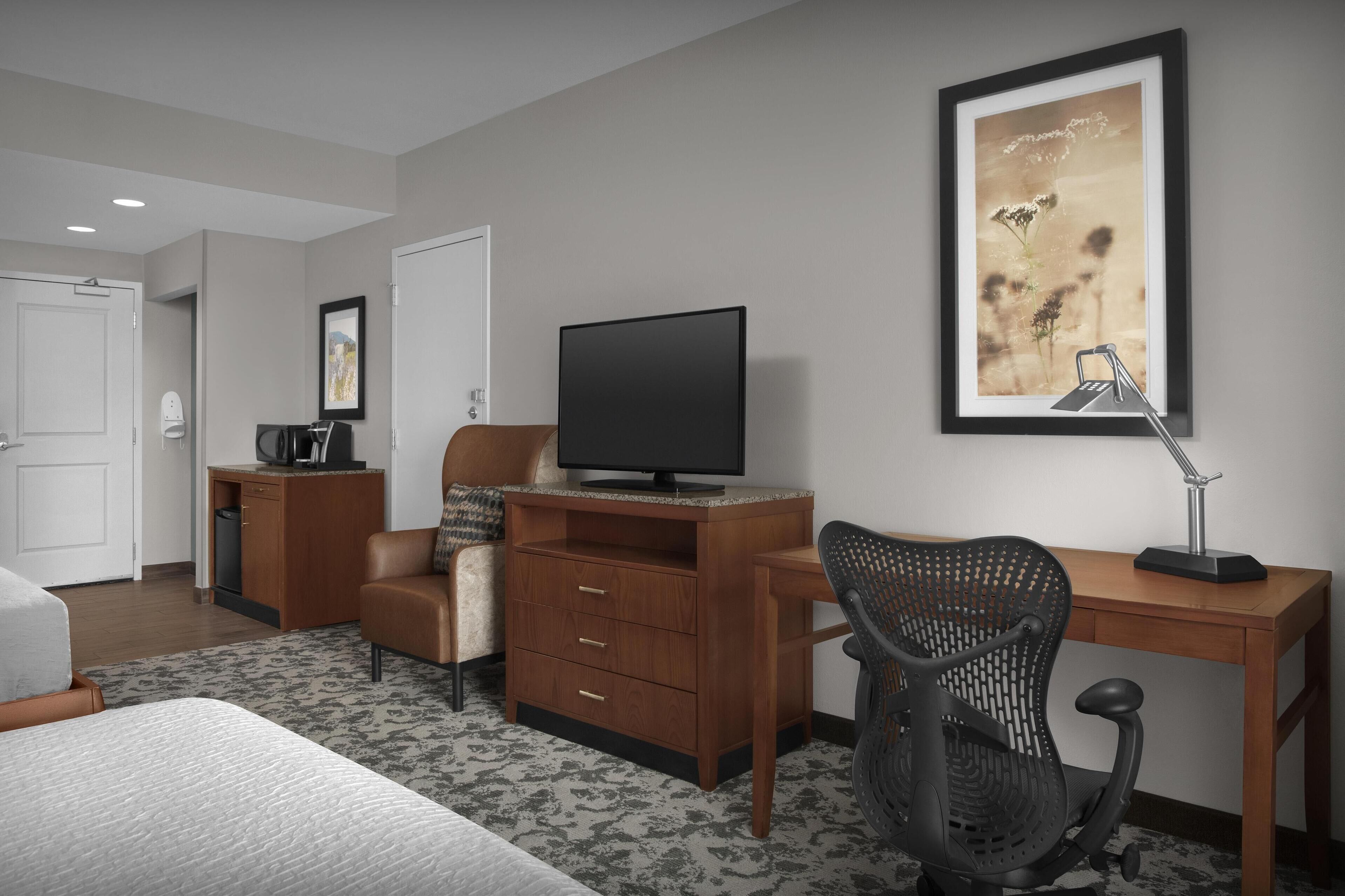 A hotel room with a bed, a desk with a computer, a chair, a television, and a framed picture on the wall.