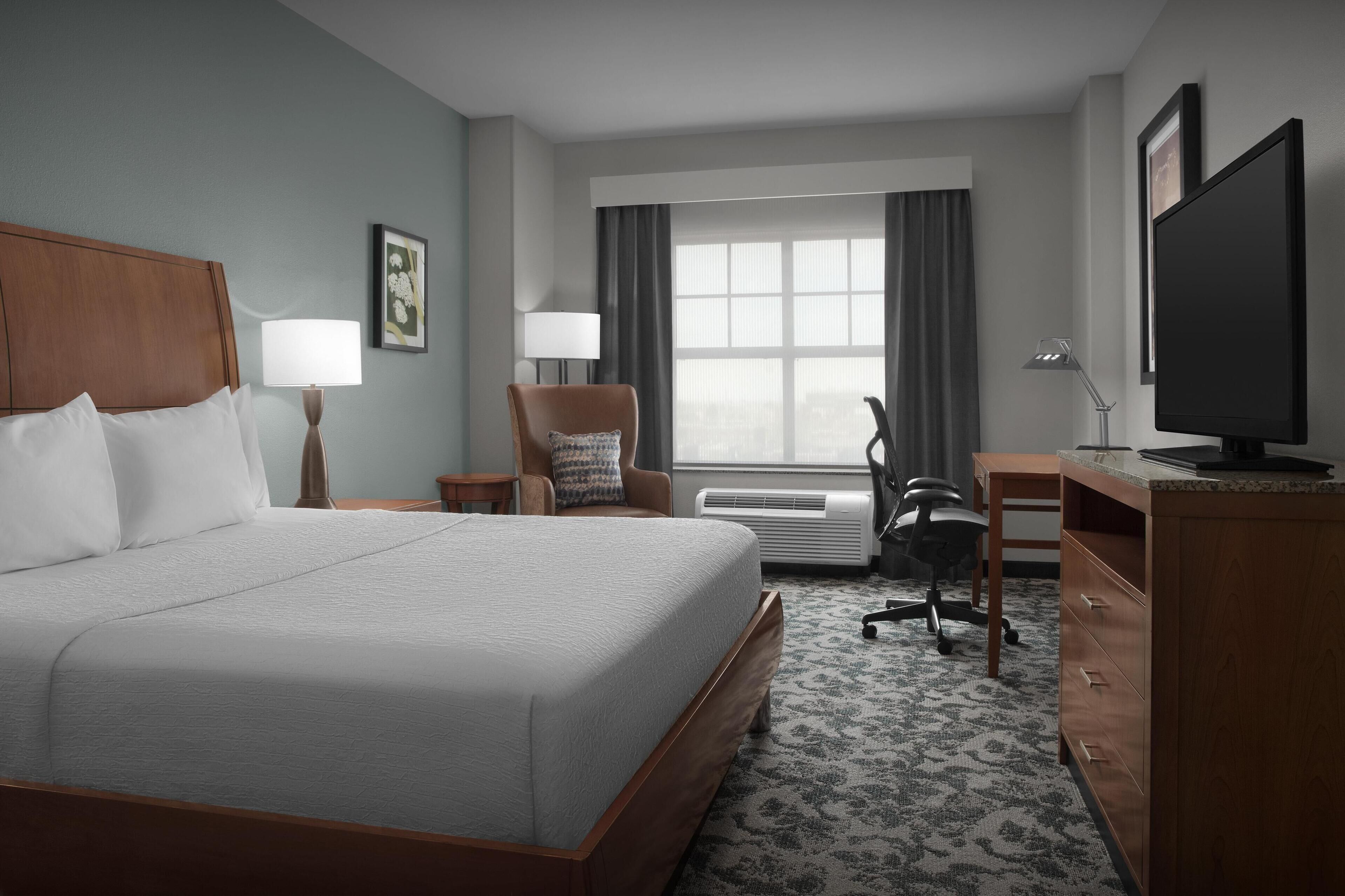 A hotel room with a large bed, a desk, a chair, a television, and a window with curtains.