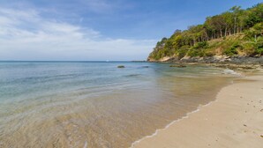 Une plage à l’eau claire, avec un rivage sablonneux et une colline boisée.