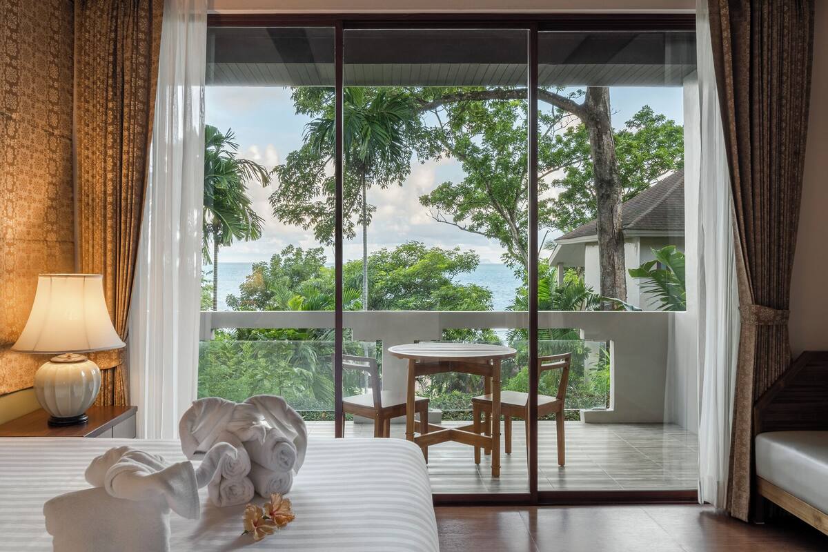 A hotel room with a bed, a table with chairs, and a balcony with a view of trees and the sea.