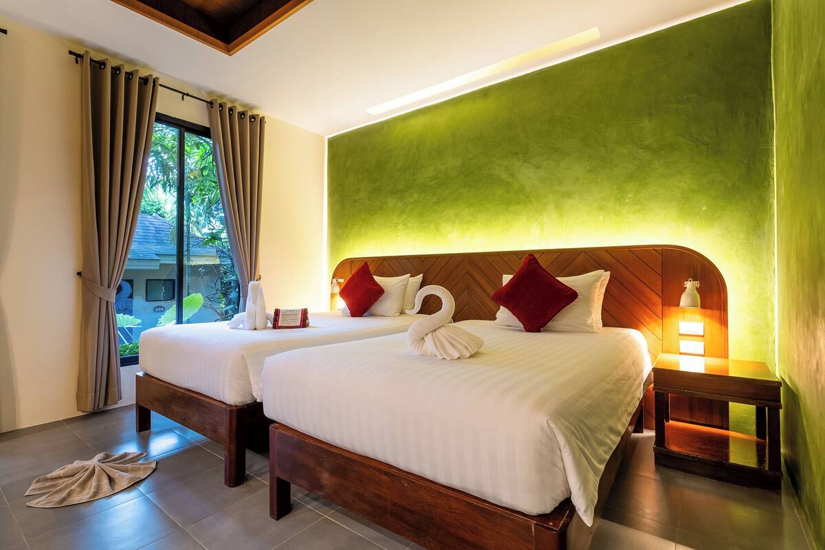 A hotel room with two beds, a green accent wall, and a view of greenery outside.