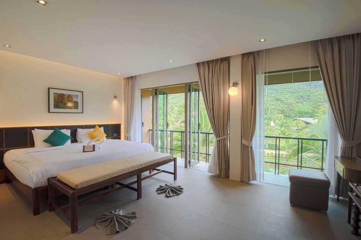 A modern hotel room with a large bed, a bench, and a balcony with a view of greenery.