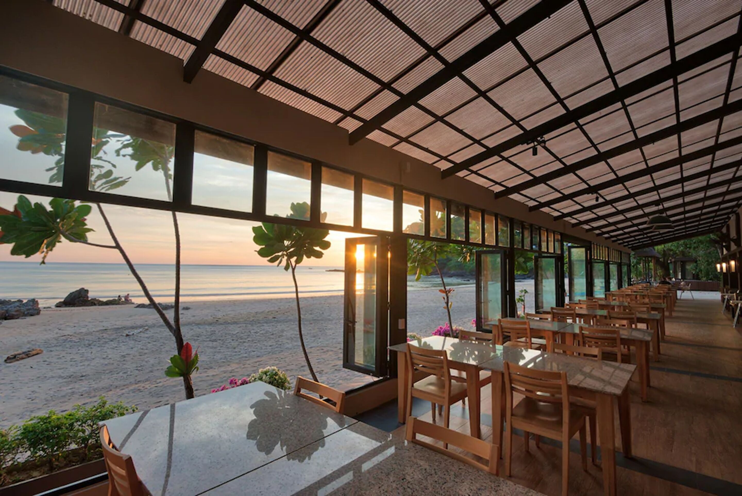A restaurant with a view of the beach, large windows, and wooden tables and chairs.