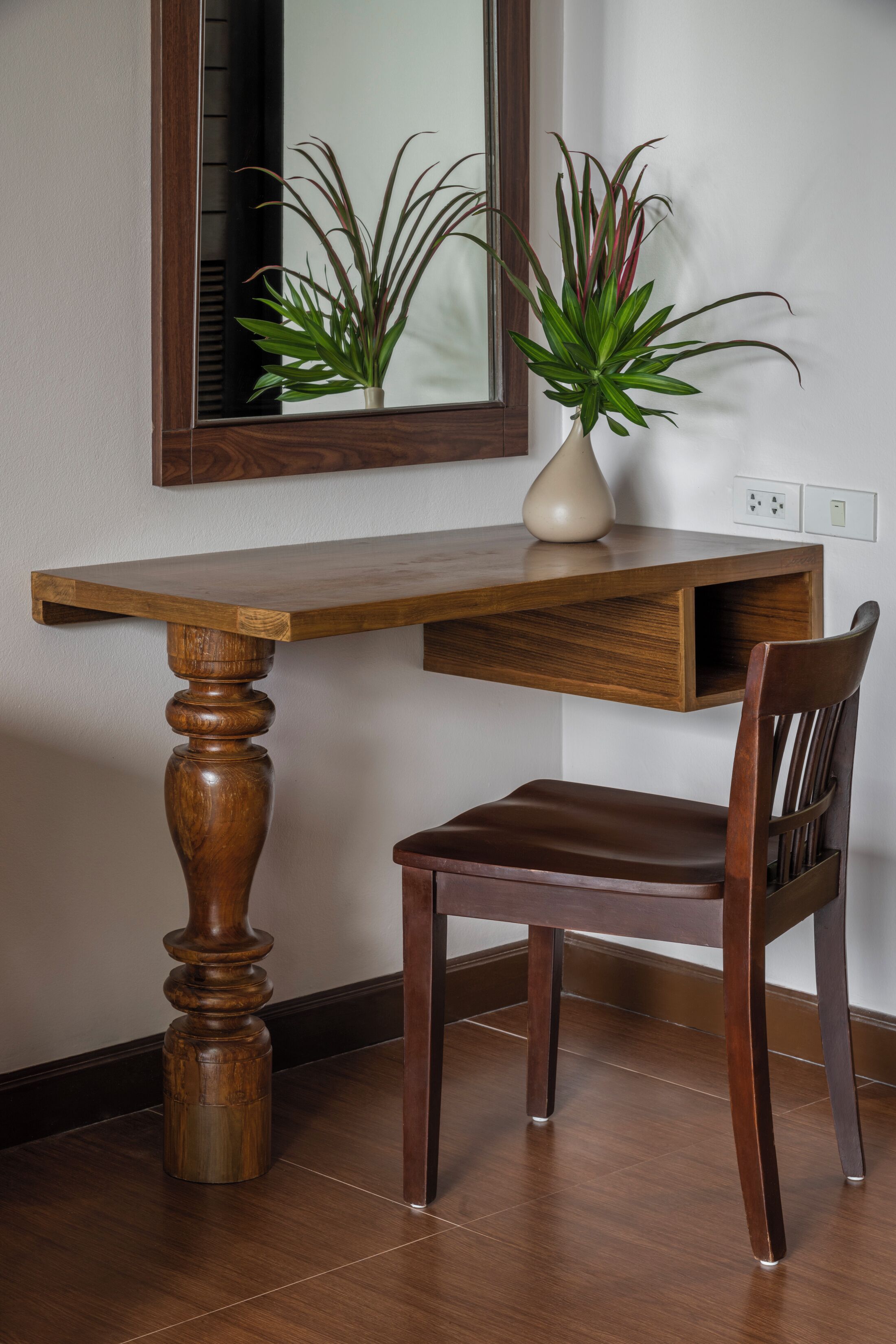 A wooden desk with a mirror above it, a vase with a plant, and a chair.