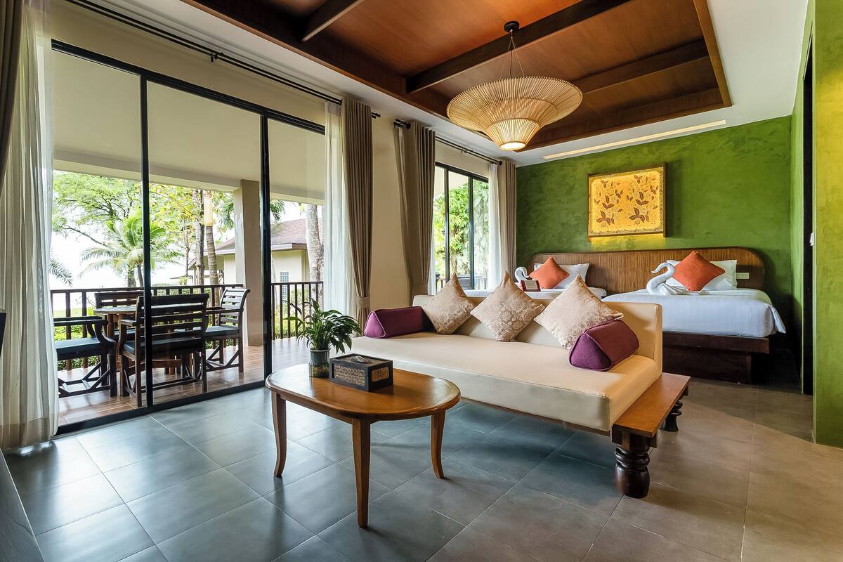 A hotel room with a sofa, a bed, a wooden coffee table, and a balcony with outdoor seating.