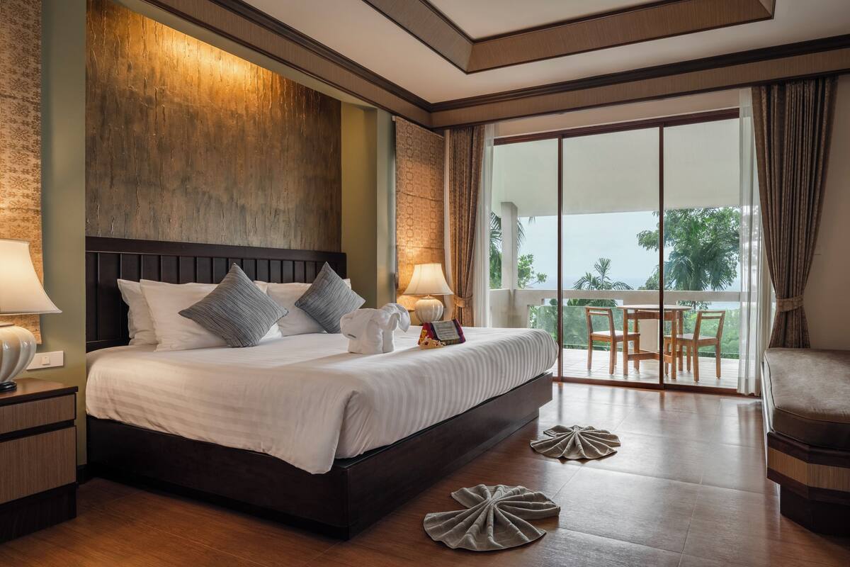 A spacious bedroom with a large bed, a balcony with a table and chairs, and a view of the outdoors.