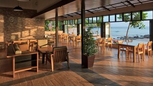 Un vaste espace intérieur doté d’un plancher en bois, de grandes fenêtres donnant sur la plage, ainsi que de plusieurs tables et chaises en bois.