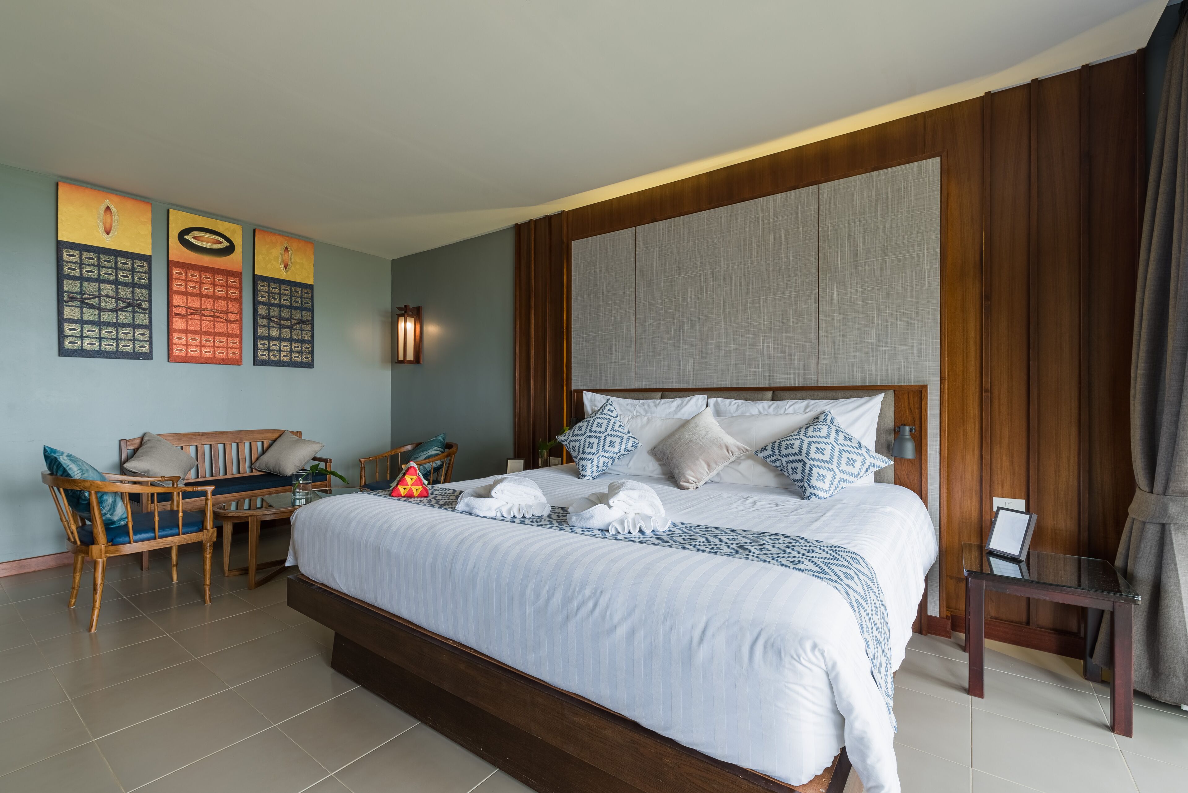 A hotel room with a large bed, a wooden headboard, a seating area with a chair and table, and a wall-mounted artwork.