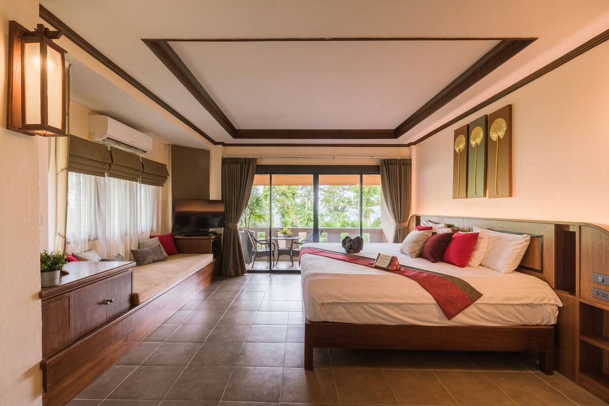 A hotel room with a large bed, a desk, and a balcony with a view of greenery.