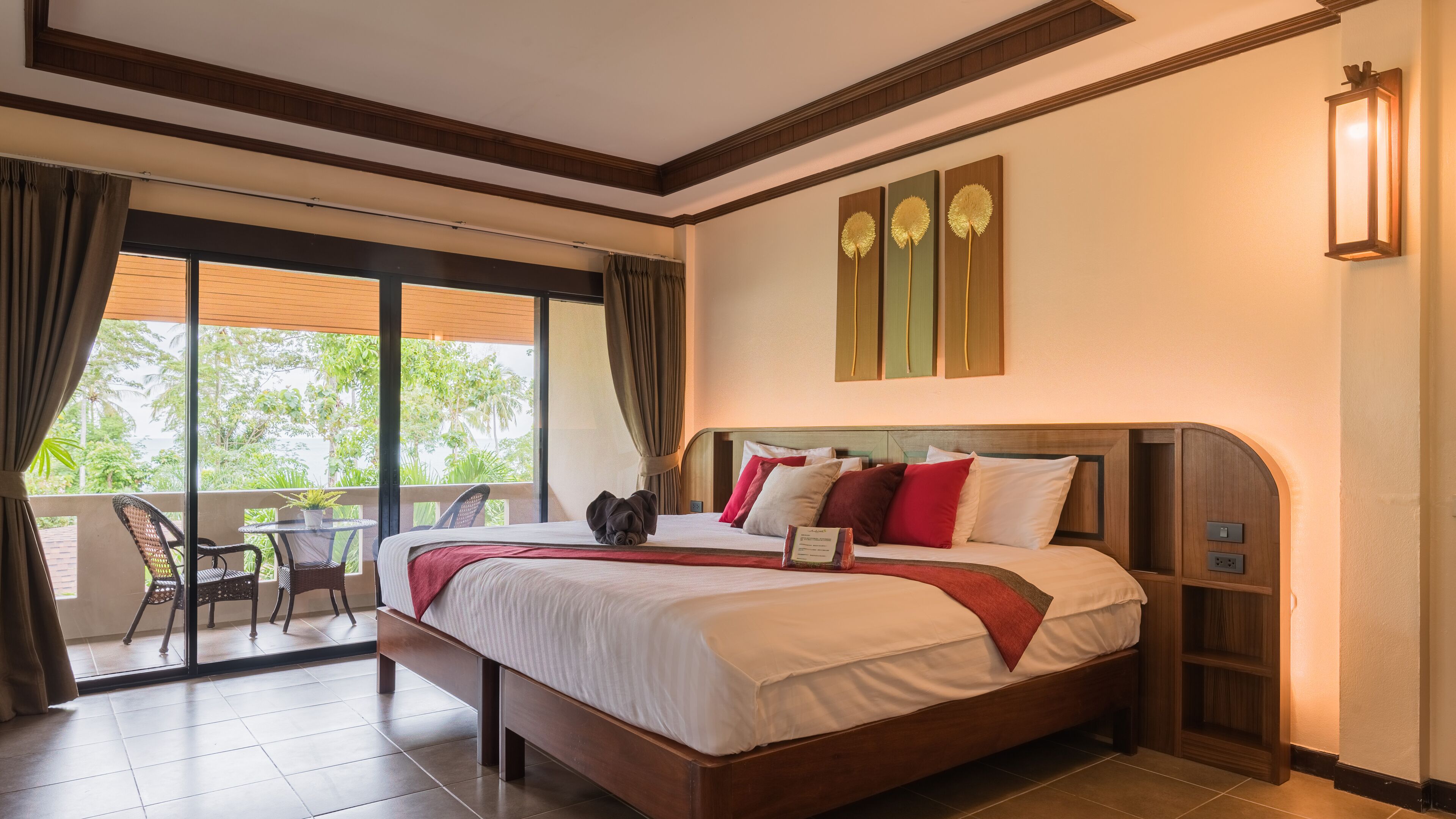 A hotel room with a large bed, a balcony with a table and chairs, and a view of greenery outside.