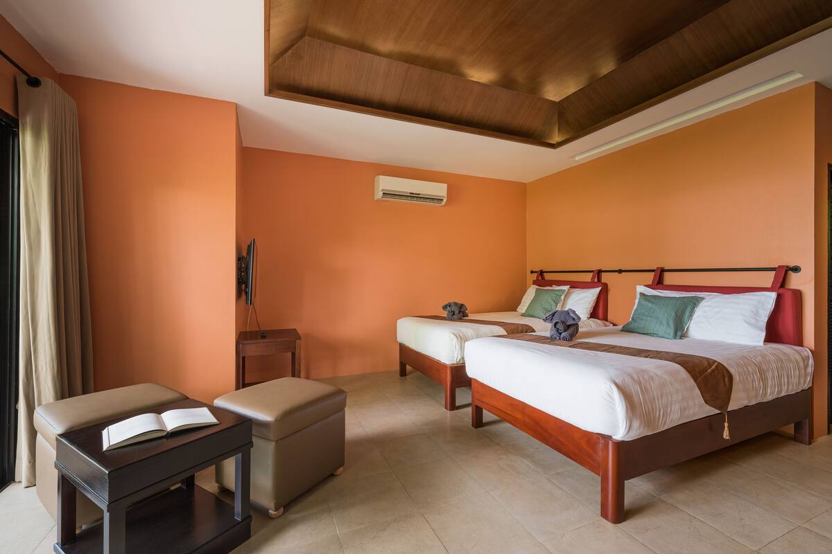 A hotel room with two beds, a small table, an ottoman, and an air conditioning unit.