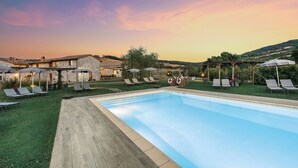 Zona junto a la piscina con sillones de descanso y sombrillas, rodeada de edificios de piedra y vistas a colinas.
