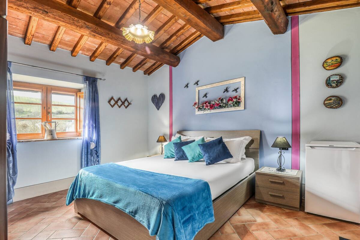 A bedroom with a wooden ceiling, a bed with a blue blanket, a nightstand with a lamp, and a wall with decorative items.
