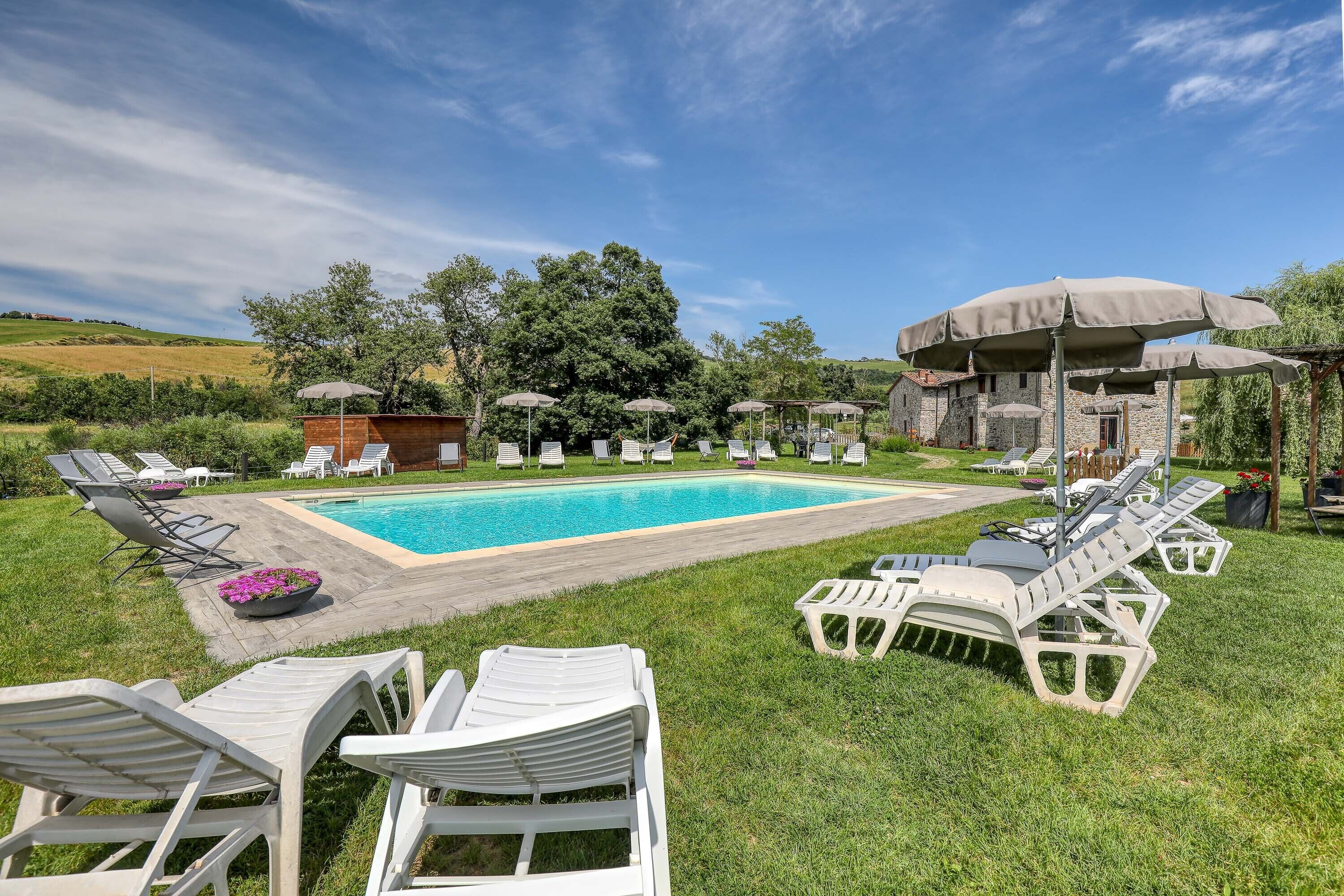 Une piscine entourée d’une pelouse et d’arbres, avec des chaises longues et des parasols à proximité.