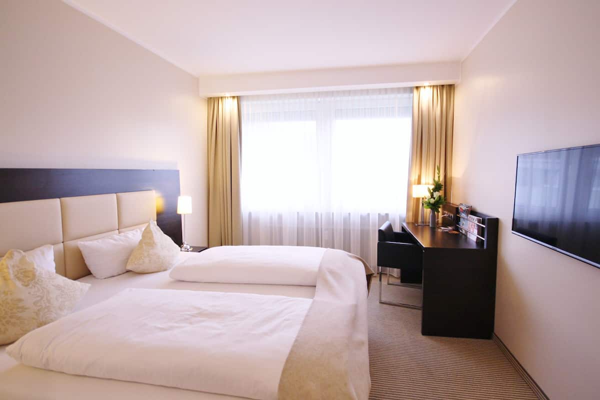Ein Hotelzimmer mit zwei Betten, einem Schreibtisch, einem Fernseher und einem Fenster mit Vorhängen.