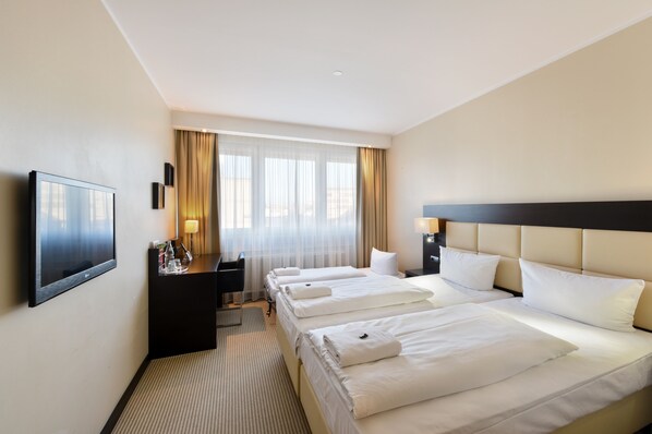 A hotel room with three beds, a desk with a chair, a TV, and a window with curtains.