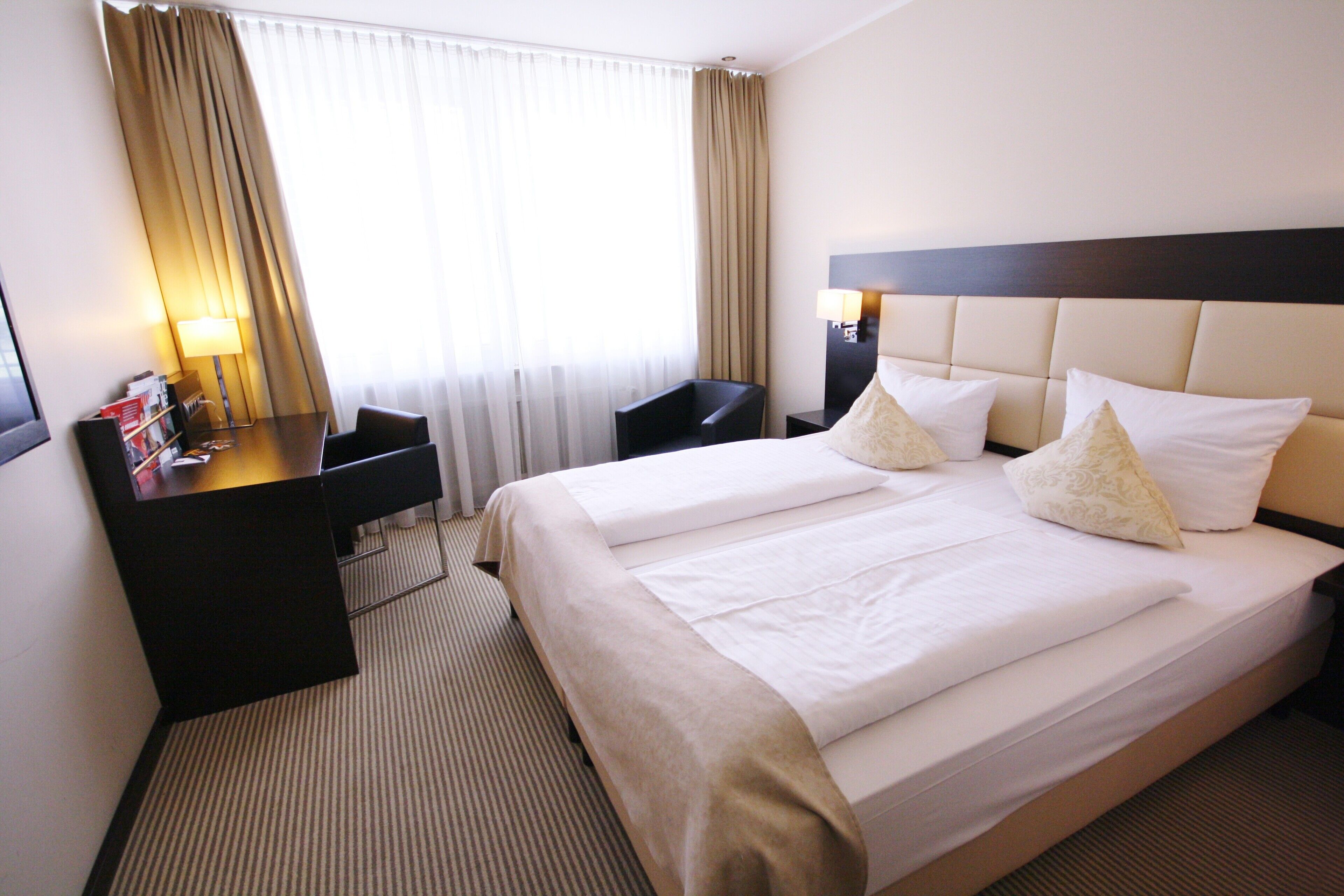 Une chambre d’hôtel avec deux lits, un bureau avec une lampe, une chaise et une grande fenêtre avec des rideaux.