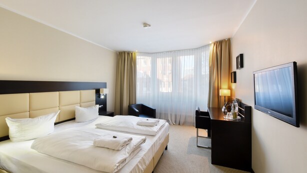 A hotel room with a large bed, a desk, a chair, a TV, and a window with curtains. - Hotel am Karlstor (Karlsruhe)