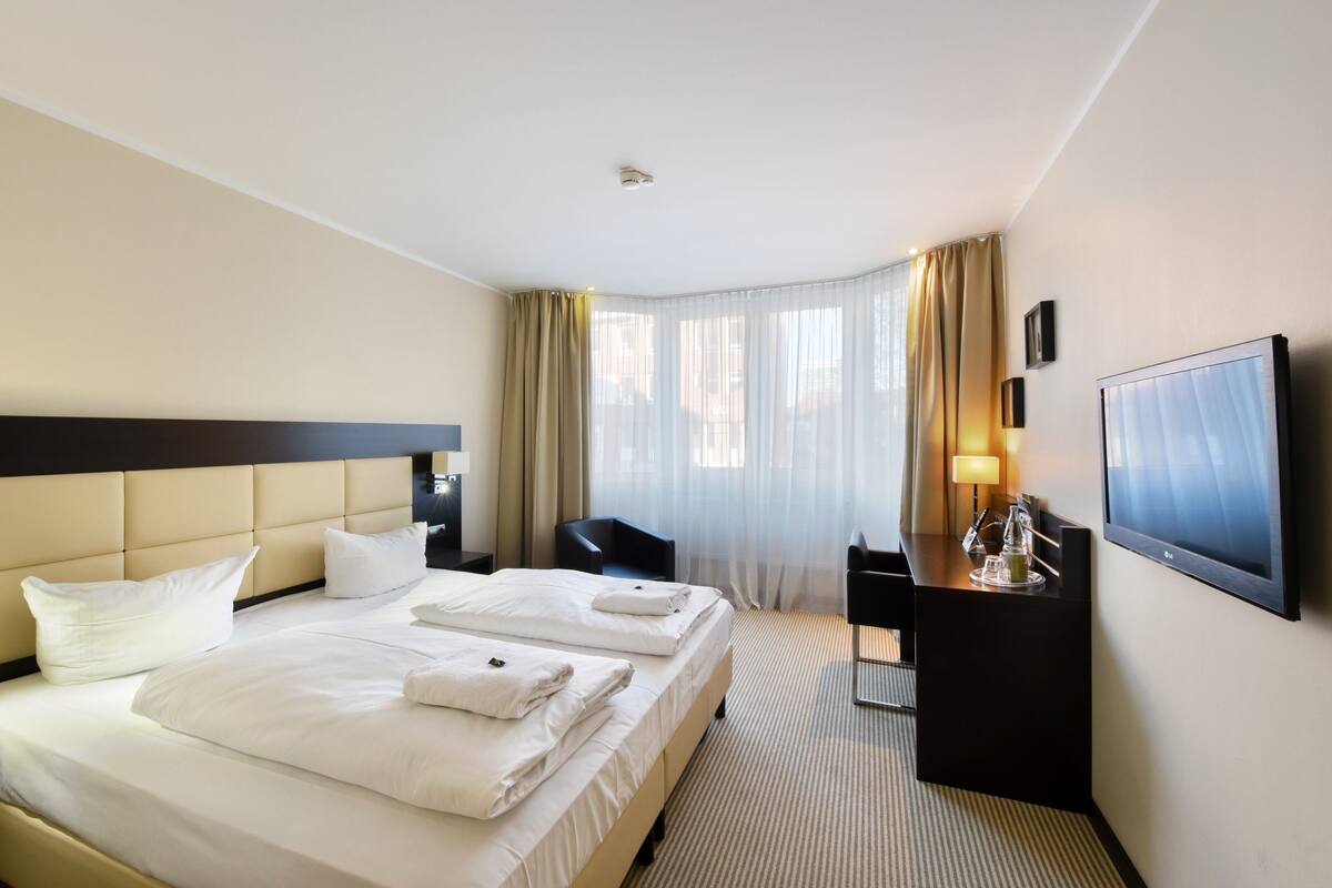 Ein Hotelzimmer mit einem großen Bett, einem Schreibtisch, einem Stuhl, einem Fernseher und einem Fenster mit Vorhängen.