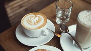 A cup of coffee with latte art on a saucer, a glass of water, and a glass of milk on a wooden table. - Hotel am Karlstor (Karlsruhe)