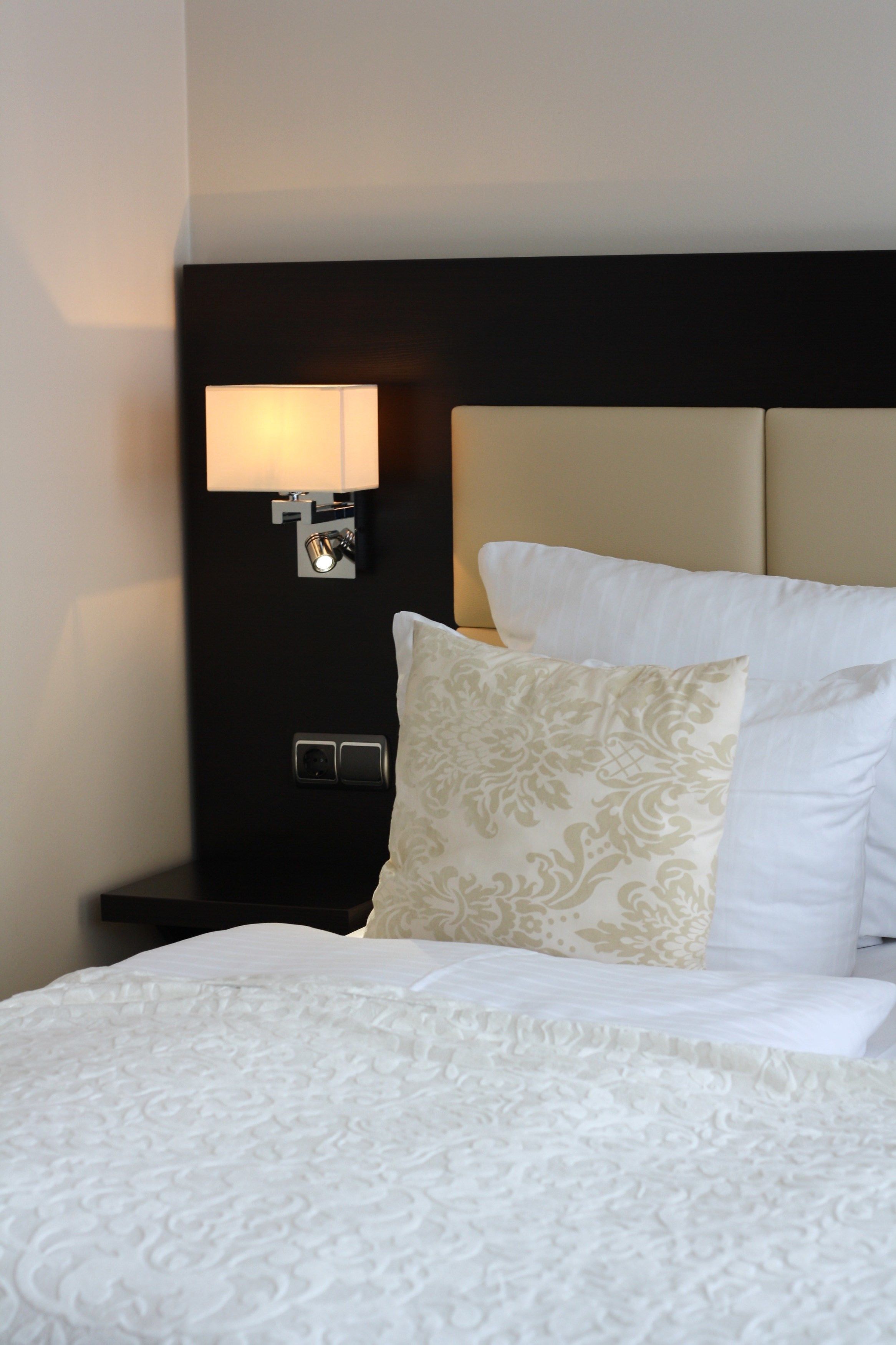 Una cama bien tendida con sábanas blancas y un cojín estampado, una lámpara de pared y un cabecero oscuro.