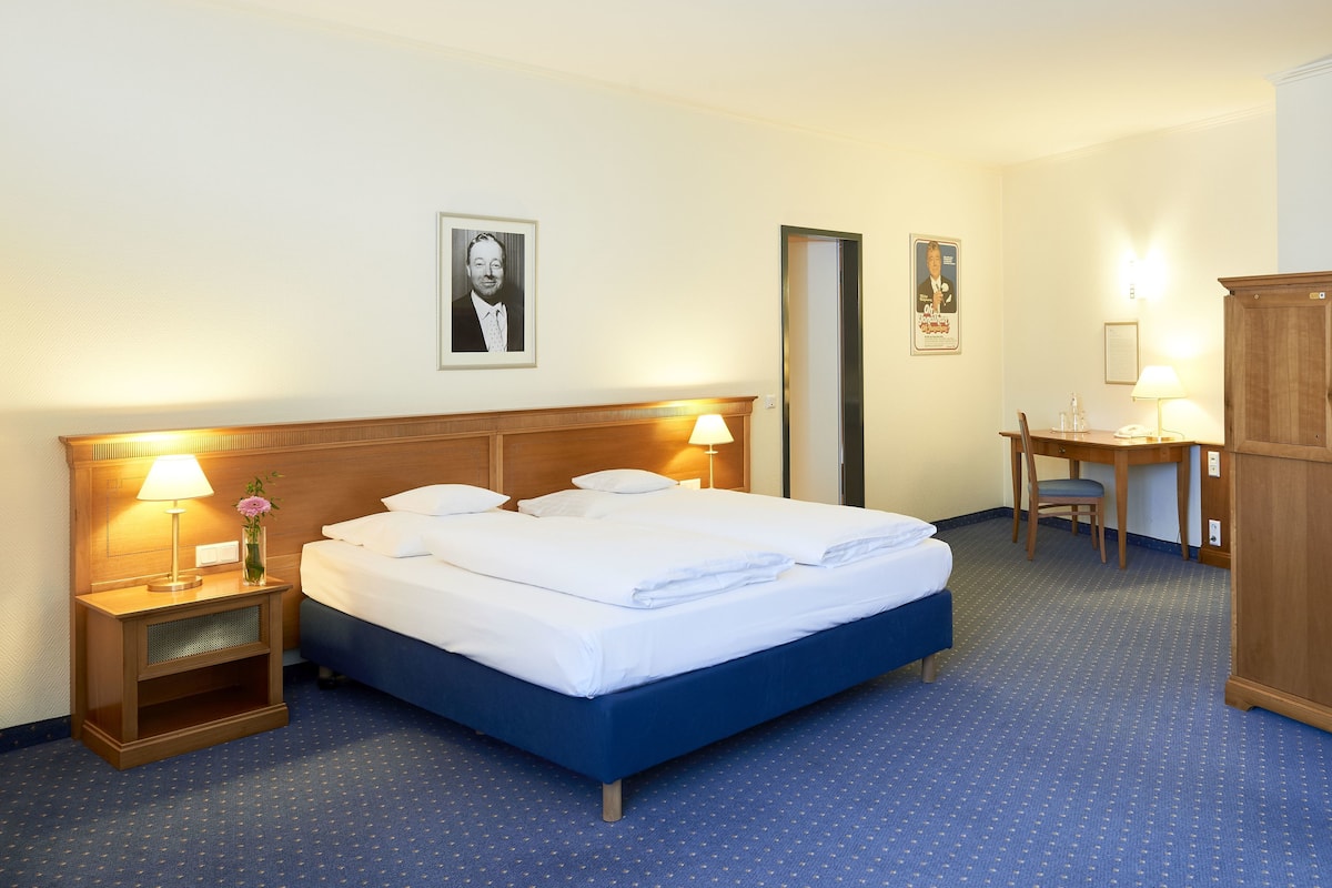 Quarto de hotel com uma cama grande, duas mesas de cabeceira com abajures, uma escrivaninha com cadeira e um guarda-roupa.