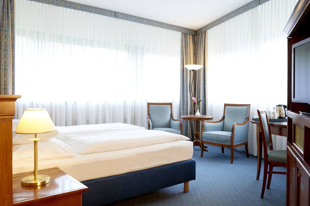 Quarto de hotel com uma cama, duas poltronas, uma mesinha com um vaso, uma luminária e uma televisão.