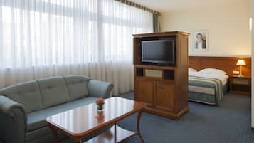 Habitación de hotel con sofá, televisor sobre un mueble de madera, cama con cabecera y mesita de noche con lámpara.