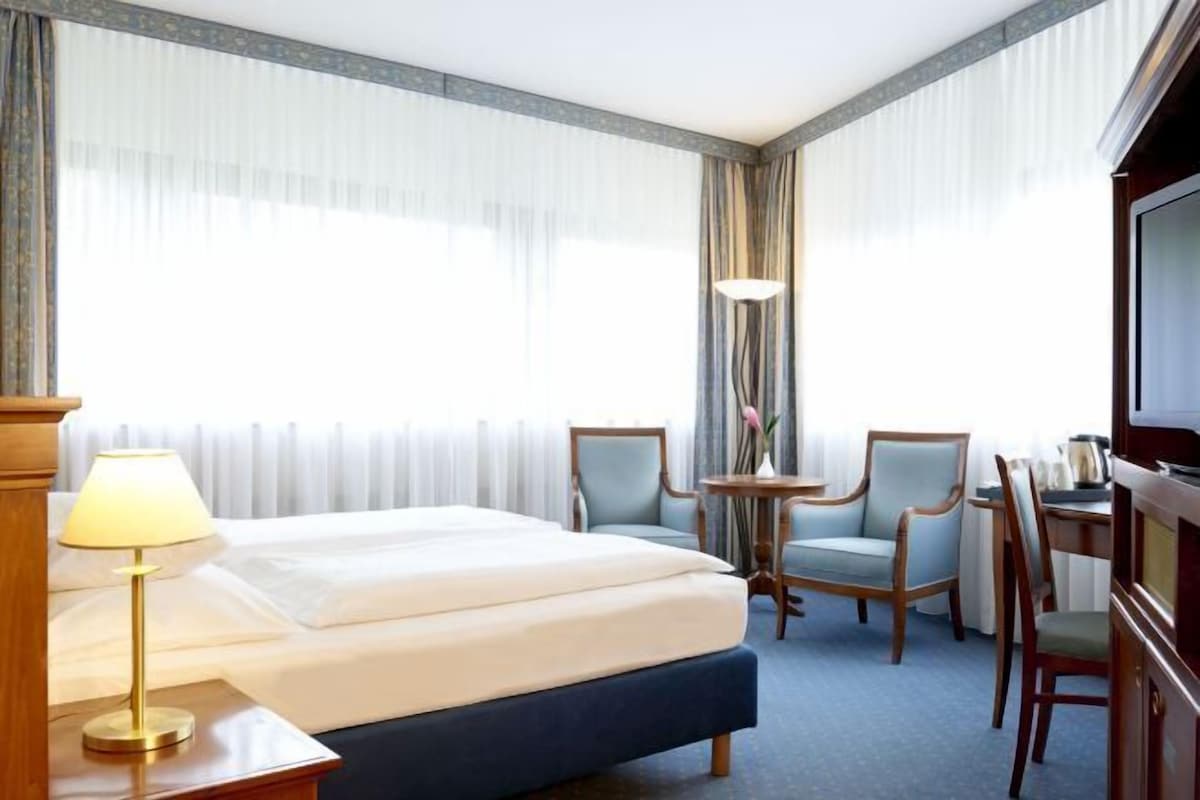 A hotel room with a bed, a desk, two chairs, a lamp, and a TV.